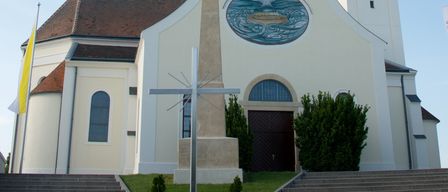 Eine weiße Kirche mit hohem Turm, Uhr und Kreuz davor. Sie hat ein braunes Dach, Glasfenster und ein großes kreisförmiges Fenster in der Mitte.