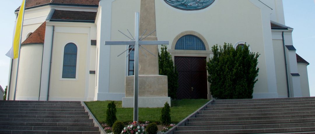 Eine weiße Kirche mit einem Turm und einer Uhr. Die Fassade zeigt ein kreisförmiges Buntglasfenster und ein Kreuz davor. Stufen führen zum Eingang, und eine Fahne befindet sich links.
