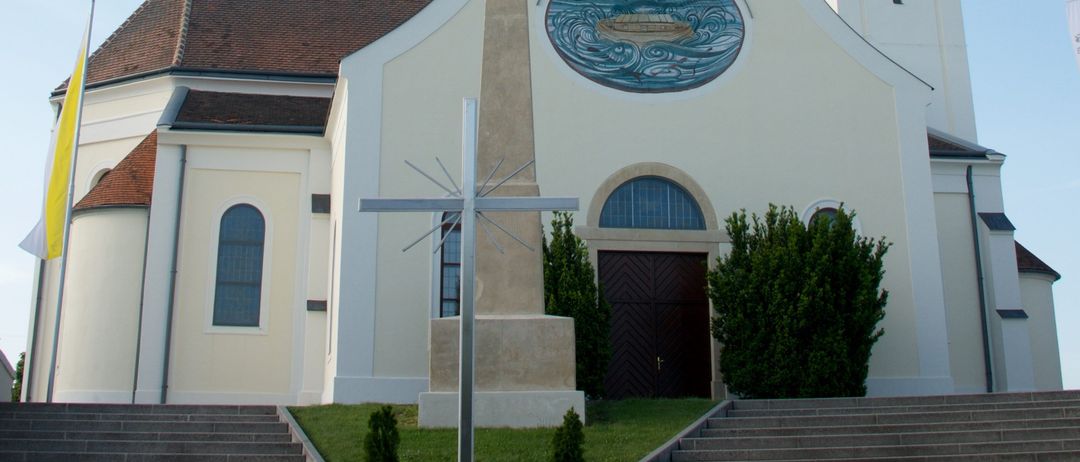 Eine weiße Kirche mit einem Turm, einer Uhr und einem Kreuz. Sie hat ein rundes Fenster und eine Statue davor. Die Kirche hat ein braunes Dach, eine Flagge auf der linken Seite und Stufen, die zu ihr hinaufführen.