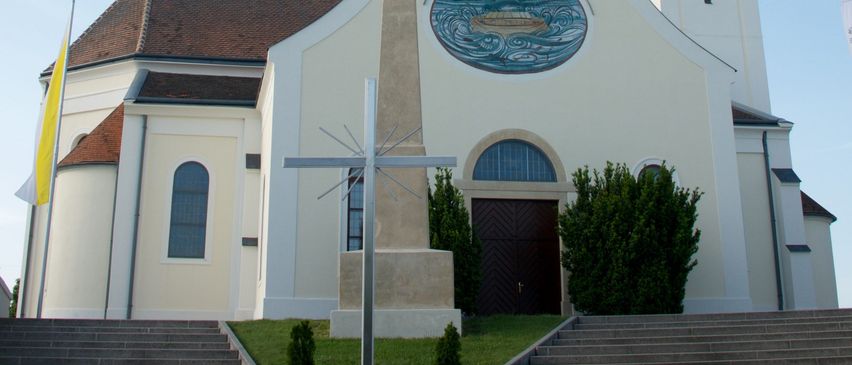 Eine weiße Kirche mit einem Turm, einer Uhr und einem Kreuz. Sie hat ein rundes Fenster und eine Statue davor. Die Kirche hat ein braunes Dach, eine Flagge auf der linken Seite und Stufen, die zu ihr hinaufführen.