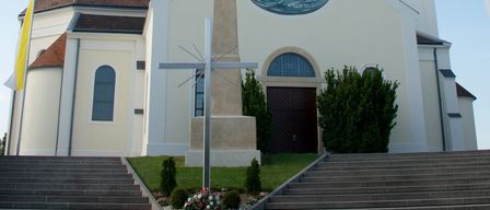 Eine weiße Kirche mit einem braunen Dach, einem Turm und einem runden Buntglasfenster. Ein Kreuz steht vor dem Eingang, mit Stufen, die hinaufführen.