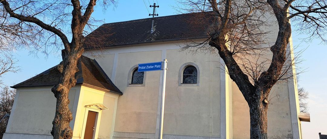 Eine Kirche mit einem Turm und einem blauen Straßenschild, umgeben von zwei kahlen Bäumen an einem sonnigen Tag.