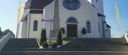 A large white church with a steeple and cross on top, featuring a clock and a circular window. It has a cross monument and flags outside.