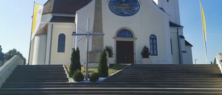 A white church with a steeple, clock, and arched window. Flags and plants are near the entrance.