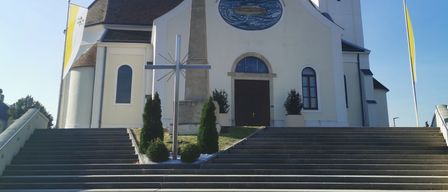 Eine weiße Kirche mit Glockenturm steht an einem sonnigen Tag. Sie hat ein Kreuz auf der linken Seite, einen Fahnenmast und ein Kreuz davor. Der Eingang hat eine Holztür.
