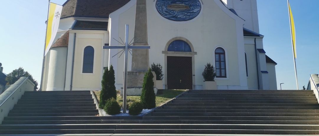 Eine weiße Kirche mit Glockenturm steht an einem sonnigen Tag. Sie hat ein Kreuz auf der linken Seite, einen Fahnenmast und ein Kreuz davor. Der Eingang hat eine Holztür.