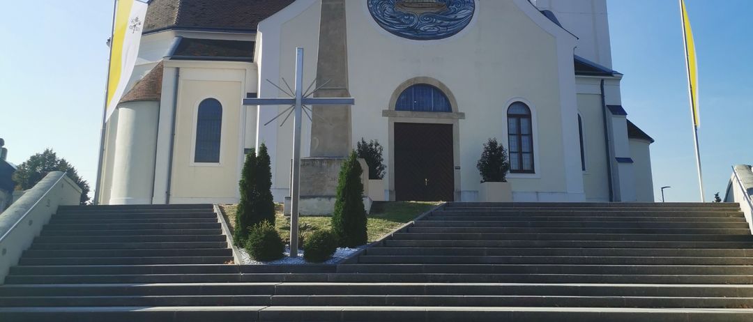 Eine große weiße Kirche mit Glockenturm, einem Kreuz auf dem Dach und einem Kreuzmonument davor unter blauem Himmel.