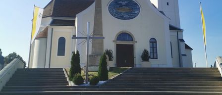 Eine weiße Kirche mit einem Turm und einer Uhr zeigt sich einem sonnigen Himmel zu. Ein Kreuz steht davor und zwei Fahnen sind auf Stangen gehisst.
