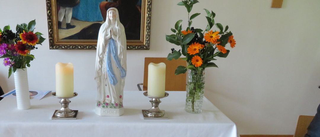 Ein Tisch mit einer weißen Tischdecke zeigt eine Statue, zwei Kerzen und zwei Vasen mit Blumen auf einem Holzboden. Ein gerahmtes Gemälde befindet sich an der Wand.