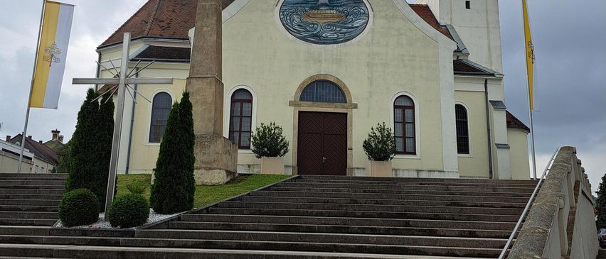 A traditional church with a steeple, a cross on top, and a clock. It has a circular stained glass window with a boat and waves. Two flags are on poles to the sides of the church.