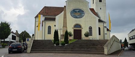 A church with a steeple, a clock, and a large window stands on a street with parked cars. The church has a large front staircase and a flag on a pole.
