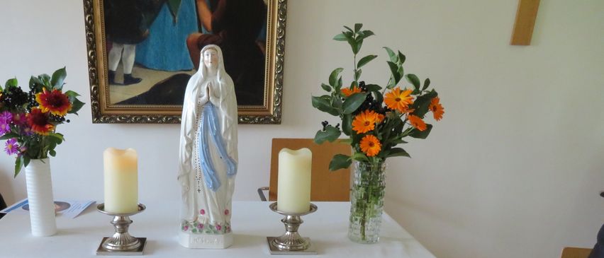 Ein weißer Altartisch, der mit einem weißen Tuch bedeckt ist. Darauf befinden sich eine Statue der Jungfrau Maria, zwei brennende Kerzen und zwei Vasen mit Blumen. Dahinter hängt ein gerahmtes Bild an der Wand.