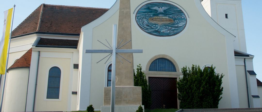 Eine weiße Kirche mit einem braunen Dach und einem hohen Turm mit einer Uhr. Die Vorderseite zeigt ein großes Kreuz und ein kreisförmiges Buntglasfenster mit einer Bootsdarstellung.