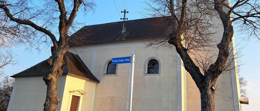 Eine Kirche mit einem Kirchturm, einem Straßenschild mit der Aufschrift Pralat Zister Platz und zwei laublosen Bäumen davor.