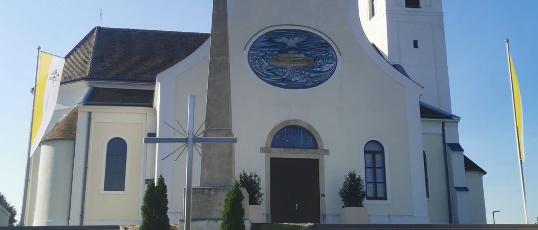 Eine weiße Kirche mit Turm und Uhren steht unter blauem Himmel. Die Kirche hat ein rundes Buntglasfenster und einen Kreuz vor sich. Zwei Fahnen wehen im Wind.