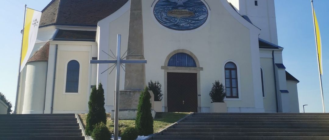 Eine weiße Kirche mit Turm, Uhr und Kreuz davor, vor einem blauen Himmel. Sie hat ein kreisförmiges Fenster mit einem blauen Design und Flaggen auf beiden Seiten.