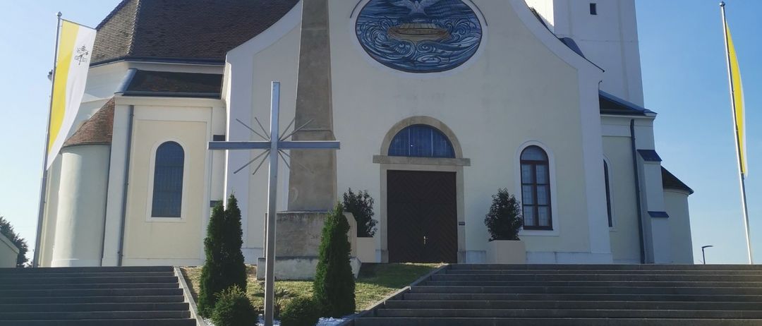 Eine weiße Kirche mit einem Turm und einem Kreuz davor, einer Uhr auf dem Turm, einem Kreuz an der Wand, einer Flagge auf der linken Seite und einer Treppe, die zu ihr hinaufführt.