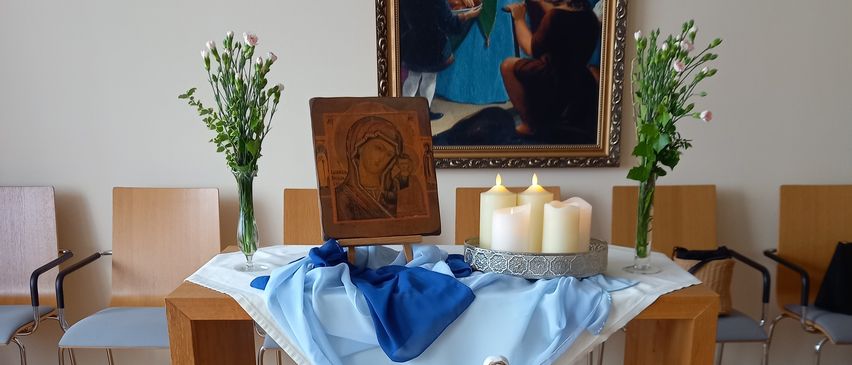 Ein Holztisch mit einem weißen Tuch darauf. Er hält eine Statue, Blumen und mehrere Kerzen. Dahinter zeigt eine Wand ein gerahmtes Gemälde.