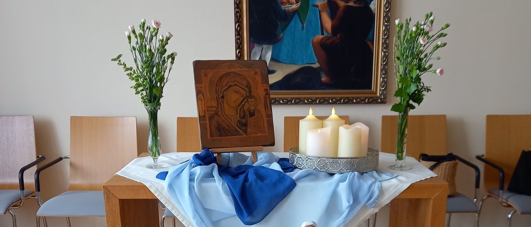 Ein Holztisch mit einem weißen Tuch darauf. Er hält eine Statue, Blumen und mehrere Kerzen. Dahinter zeigt eine Wand ein gerahmtes Gemälde.