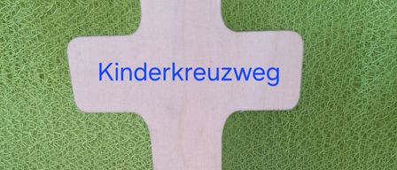 Ein hölzernes Kreuz mit dem Text 'Kinderkreuzweg' auf grünem Hintergrund, begleitet von einer gelben Blume und grünen Blättern.
