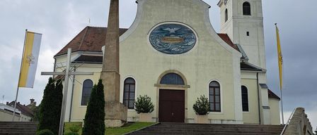 Eine traditionelle Kirche mit einem braunen Dach und einem Glockenturm. Sie hat ein rundes Fenster und einen Rundbogeneingang. Zwei Fahnen befinden sich an den Seiten.