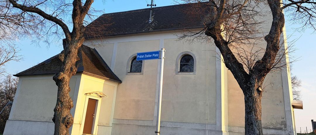 A white church with a steeple stands between two trees. A blue street sign reading 'Pralat Zistler Platz' is in front.