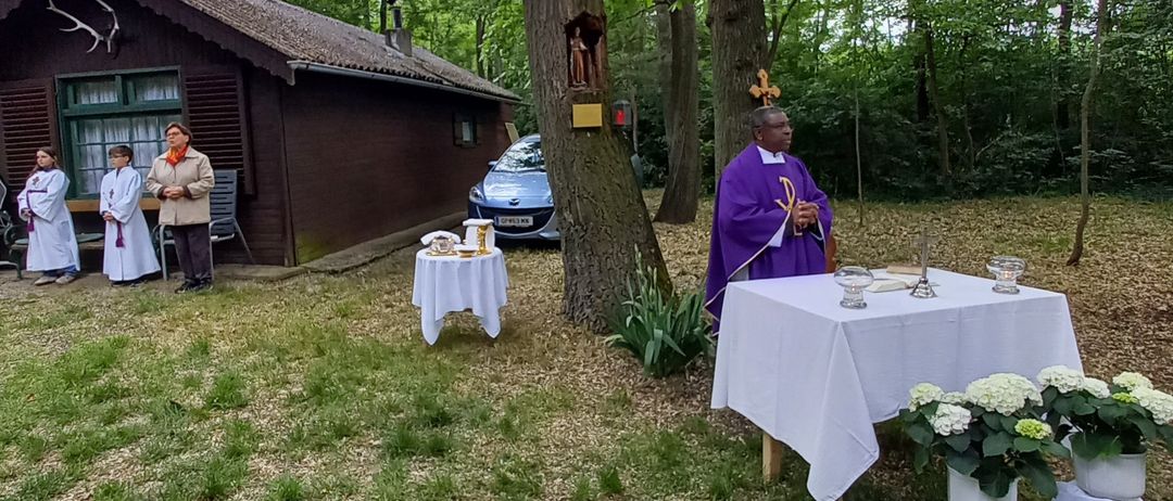 A priest stands beside a table with candles in a forest clearing, with a small house and a car visible in the background.