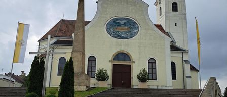 A traditional church with a pointed tower, a large round painting, and a cross in front. The church has flags on both sides and steps leading up to it.