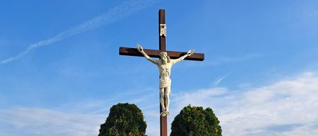 A large wooden cross with a statue of Jesus stands in a vineyard. It is flanked by two trees and a bench. In the distance, wind turbines and a clear blue sky are visible.