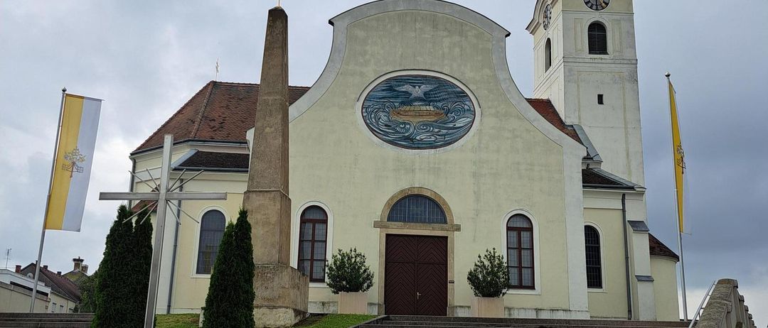 Eine Kirche mit Turm und Dachreiter steht mit einem kreisförmigen Wandbild, Fahnen und einem Kreuz davor. Sie hat Stufen zum Eingang, umgeben von Pflanzen und Gras.