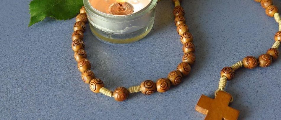Eine hölzerne Rosenkranz, eine brennende Kerze und eine gelbe Rose auf einem grauen Hintergrund. Die Kerze ist in einem kleinen Glasbecher.