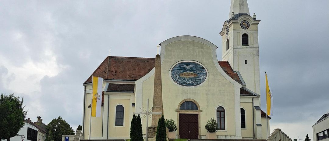 Eine Kirche mit einem Turm und einem Glockenturm steht an einer Straße mit geparkten Autos davor. Die Kirche hat ein großes Fenster und eine Treppe, die zu ihrem Eingang führt.