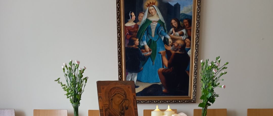 A wooden table covered with a blue cloth and adorned with a statue, flowers, and candles. Behind it, a painting and more flowers. Chairs are arranged around.