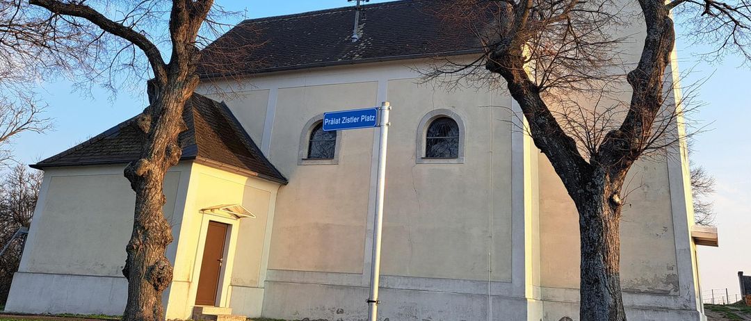 A church with a tower, surrounded by leafless trees, is seen under a clear blue sky. It has a brown roof and a sign reading Pralat Zistler Platz.