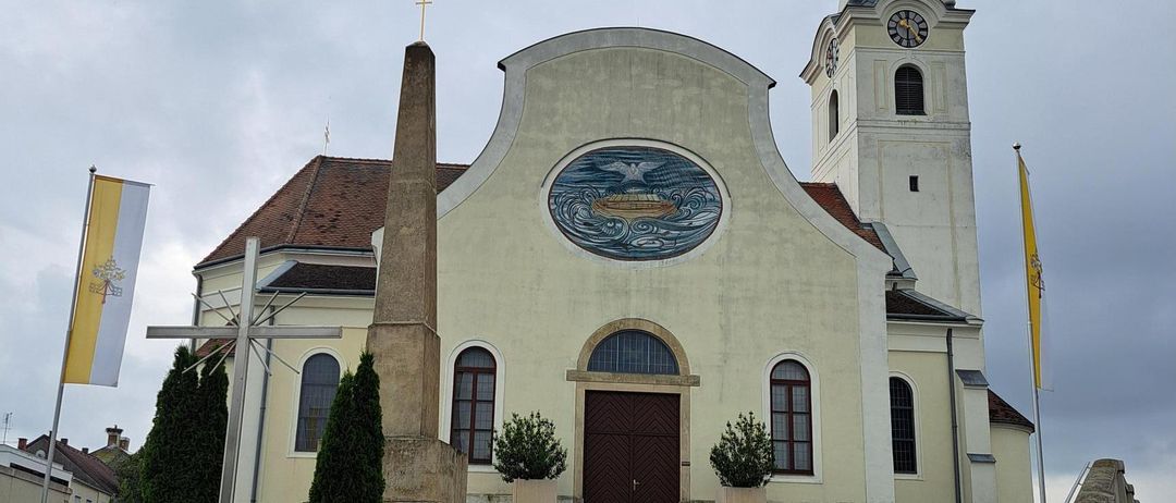 A traditional church with a clock tower and a large window. Flags and a cross adorn the front.