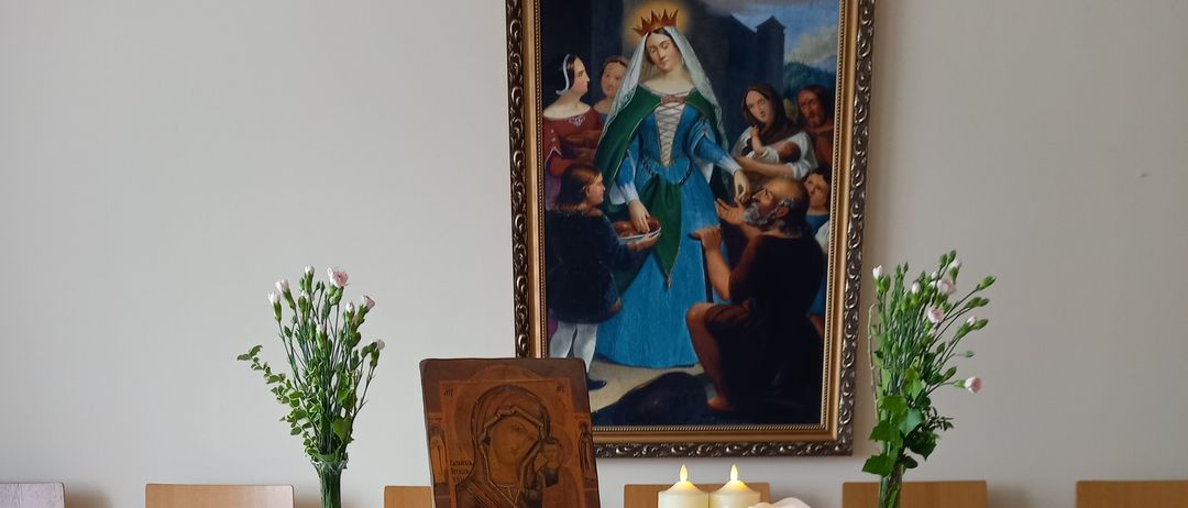 A table with a white cloth, a statue of Mary, two candles, and flowers. A painting hangs on the wall. Chairs surround the table.