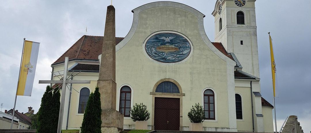 Eine weiße Kirche mit einem Turm, der eine Uhr und zwei Kreuze auf der Spitze hat. Sie verfügt über ein rundes Buntglasfenster und ein Kreuz davor. Es gibt zwei Fahnen und eine große Statue links.