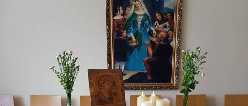 A wooden table holds a small statue of Mary, flanked by two candles. The table is covered in white and blue fabric. There are two vases of flowers and a framed religious image behind the table.