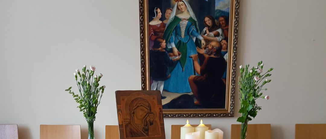 Ein hölzerner Tisch trägt eine kleine Statue von Maria, flankiert von zwei Kerzen. Der Tisch ist mit weißer und blauer Stoff bedeckt. Hinter dem Tisch befinden sich zwei Vasen mit Blumen und ein gerahmtes religiöses Bild.