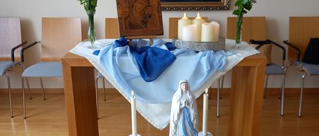 Ein Altar mit einer Statue der Jungfrau Maria, Kerzen, Blumen und einem Gemälde an der Wand. Die Umgebung umfasst Stühle und einen Holztisch.