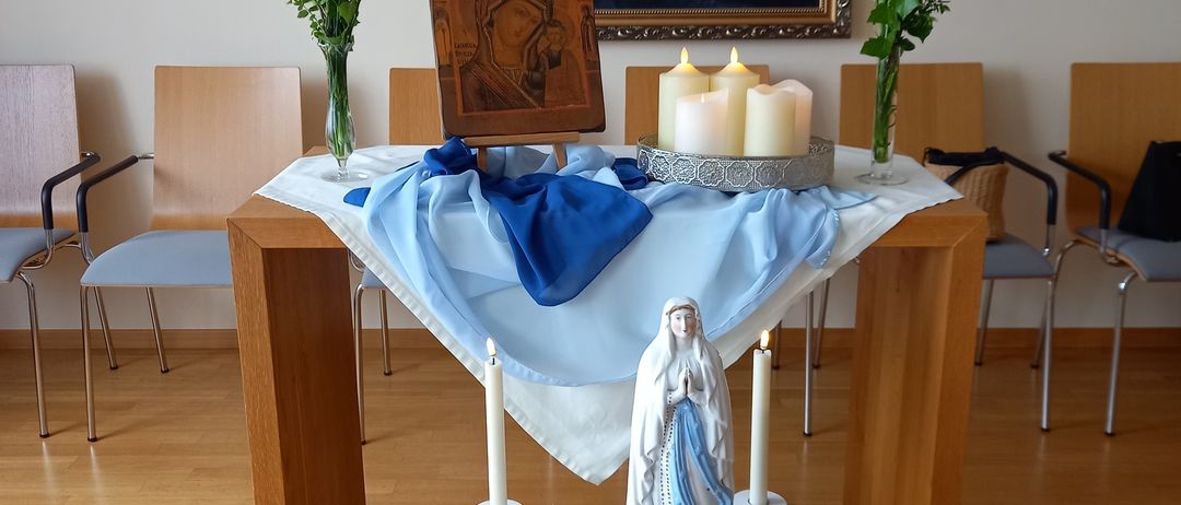 Ein Altar mit einer Statue der Jungfrau Maria, Kerzen, Blumen und einem Gemälde an der Wand. Die Umgebung umfasst Stühle und einen Holztisch.