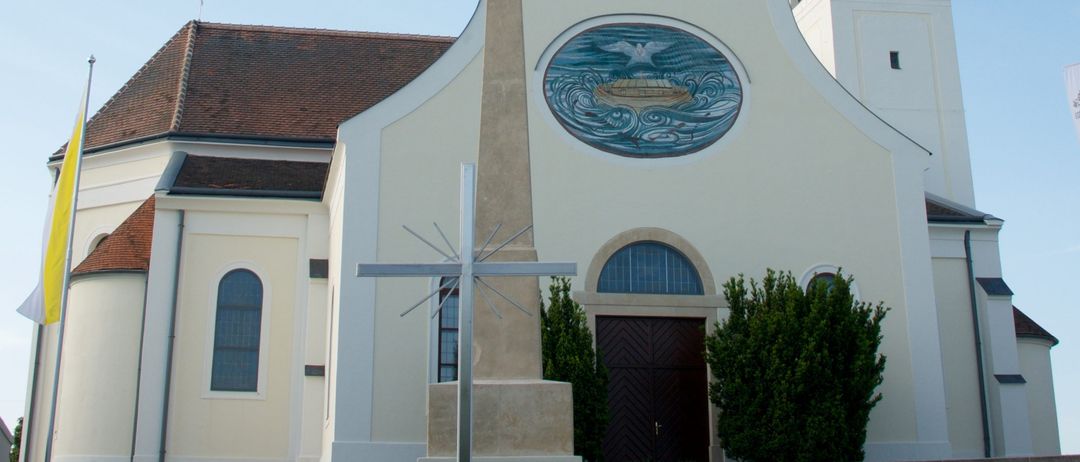 Eine weiße Kirche mit Turm und Uhr, mit einem kreisförmigen Buntglasfenster und einem Kreuz davor.