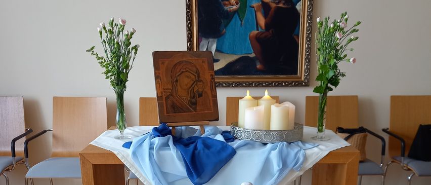 Ein hölzerner Tisch mit einem blauen Tuch, einer Statue von Maria und mehreren Kerzen. Dahinter ein Gemälde einer Familienszene. Zwei Vasen mit Blumen auf beiden Seiten.