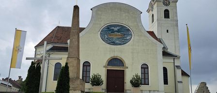 Eine Kirche mit einem Kirchturm und einem Glockenturm, mit einem großen kreisförmigen Gemälde über dem Eingang, flankiert von zwei Fahnen und einem Kreuz.