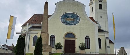 A church with a large round window, a steeple, and a clock tower. The church has a brown roof and a white wall. There are two flags on the side of the church.