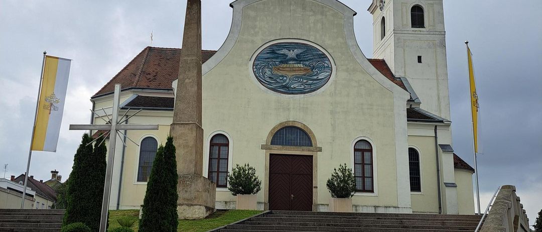 A traditional church with a steeple, a round window, and a flagpole on the left side. The church has a brown roof and is surrounded by a lawn with plants.