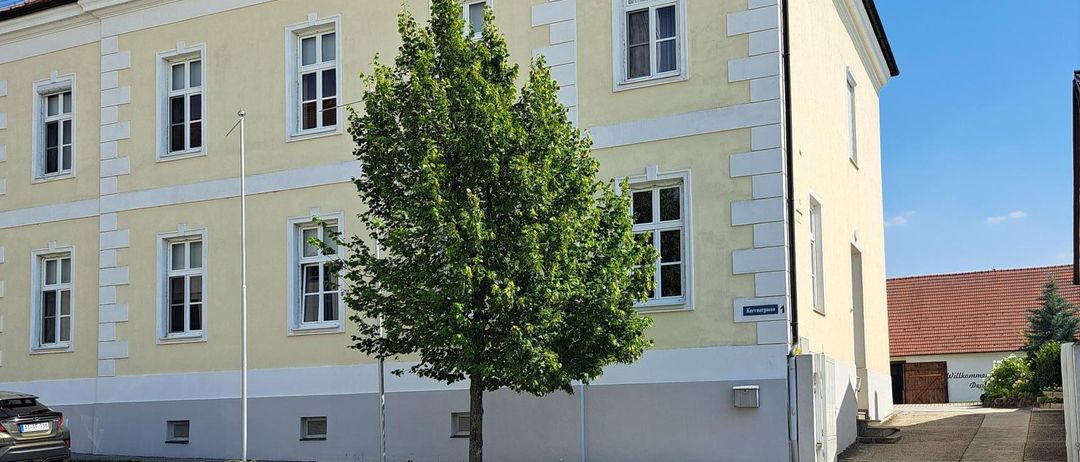 A beige building with multiple windows and a brown roof, a tree in front, and a car parked to the left.