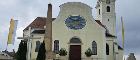 Eine Kirche mit einem Turm und einem großen kreisförmigen Emblem an ihrer Fassade. Die Kirche hat ein braunes Dach und ist von einem Rasenbereich mit Pflanzen und einem Kreuz umgeben. Auf beiden Seiten der Kirche befinden sich zwei Fahnen.