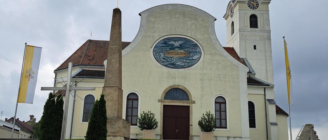 Eine Kirche mit einem Turm und einem großen kreisförmigen Emblem an ihrer Fassade. Die Kirche hat ein braunes Dach und ist von einem Rasenbereich mit Pflanzen und einem Kreuz umgeben. Auf beiden Seiten der Kirche befinden sich zwei Fahnen.