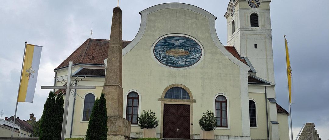 Eine traditionelle Kirche mit einem Kirchturm, einer Uhr und einem großen runden Fenster mit einem Schiffsdesign. Es hat eine Flagge auf der linken Seite und ein Kreuz auf der rechten Seite.